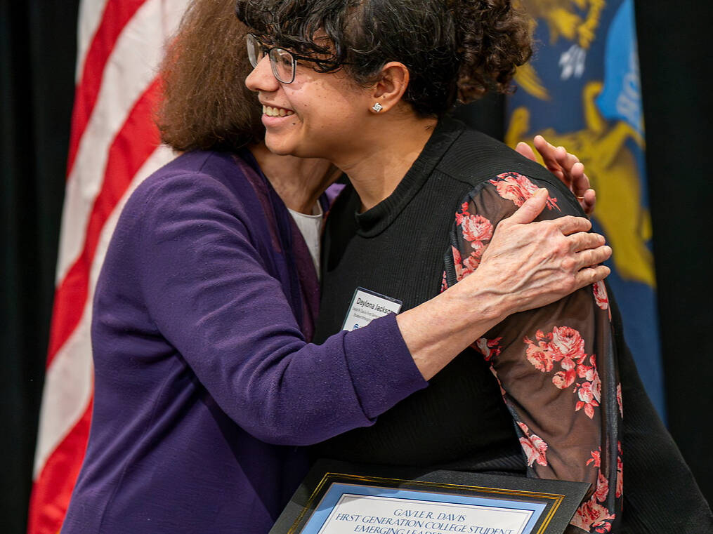 A person in a purple outfit embraces another person holding a framed award, with an American flag and a state flag visible in the background.